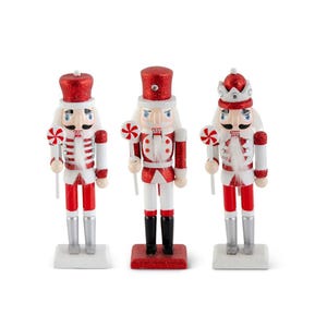 May include: Three decorative nutcracker figurines. Each figure is dressed in a red and white uniform with a matching red hat. The nutcrackers hold a lollipop and stand on a white or red base. The figurines have silver or black boots.