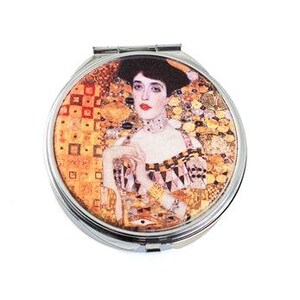 May include: A round, silver compact mirror featuring a portrait of a woman with red lips and a black hat. The artwork has a gold and orange mosaic background, reminiscent of Gustav Klimt's style. The compact is closed and has a shiny, metallic finish.