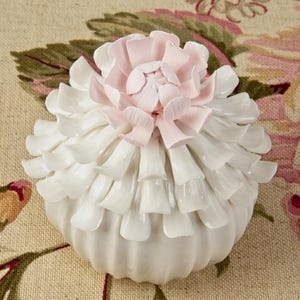 May include: A white ceramic decorative container with a flower-shaped lid. The lid is a light pink color and the container has a ruffled design. The container is sitting on a floral patterned fabric.