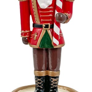 May include: A decorative Christmas nutcracker figurine. The figurine is dressed in a red uniform with gold accents, black boots, and a white beard. It holds a candy cane and stands on a drum base.