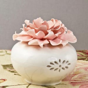 May include: A white ceramic vase with a light pink peony flower lid. The vase has a round shape with a floral design cut-out and sits on a floral patterned surface. The peony is a soft, pastel color.