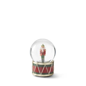 May include: A festive snow globe featuring a miniature nutcracker soldier inside a clear glass sphere. The base is designed to resemble a drum, with red and green panels and gold accents. A charming holiday decoration.