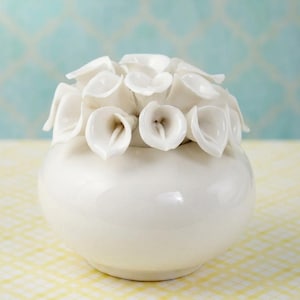 May include: A white ceramic vase with a rounded base and a cluster of white calla lily flowers on top. The vase is glossy and sits on a yellow and white checkered surface, with a light blue patterned background.