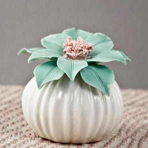 May include: A white ceramic vase with a ribbed design, topped with a large, stylized flower. The flower has light green petals and a pink center. The vase is on a textured surface.