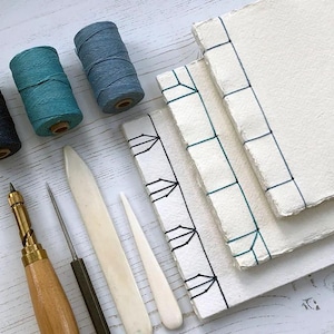 May include: Bookbinding tools and materials, including blue and black thread spools, bone folders, needles, and bound paper. The items are arranged on a light wood surface, ideal for a craft or DIY project.
