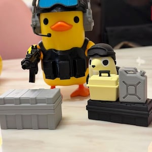 May include: A yellow duck figurine wearing a black tactical vest, headset, and holding a toy gun. A smaller duck figurine is also present, along with gray and yellow storage containers and a gray fuel canister.
