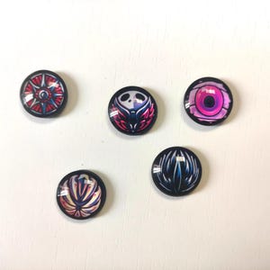 May include: Five round, glossy buttons with various colorful designs. The designs include a red and blue wheel, a skull with pink wings, a pink and purple eye, a white and yellow floral design, and a blue and black abstract design.