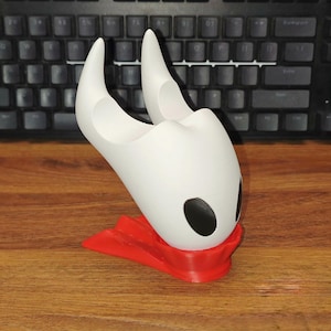 Peut inclure: Figurine blanche et rouge imprimée en 3D représentant un personnage avec une tête à cornes et des yeux noirs. La figurine est posée sur une base rouge et sur une surface en bois. Un clavier noir est en arrière-plan.