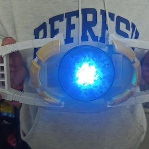 May include: A white and gold toy with a glowing blue center, held in front of a person. The toy has a rectangular frame with a circular center that emits bright blue light. The Lion Rock Toys logo is visible in the lower left corner.