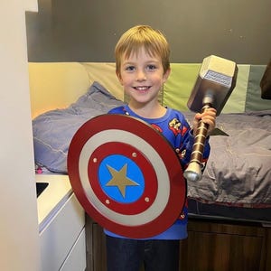 K&ouml;nnte beinhalten: Ein Kind h&auml;lt einen Captain America-Schild und Thors Hammer. Der Schild ist rot, wei&szlig; und blau mit einem goldenen Stern. Der Hammer ist silber und braun. Das Kind tr&auml;gt ein blaues Oberteil und dunkle Jeans.