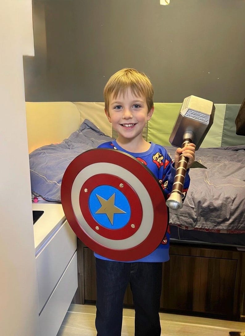 K&ouml;nnte beinhalten: Ein Kind h&auml;lt einen Captain America-Schild und Thors Hammer. Der Schild ist rot, wei&szlig; und blau mit einem goldenen Stern. Der Hammer ist silber und braun. Das Kind tr&auml;gt ein blaues Oberteil und dunkle Jeans.