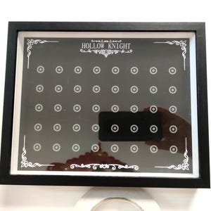 May include: A framed black and white display board with the text "HOLLOW KNIGHT" at the top. The board has a grid of small circles and decorative corner designs. A hand holding a phone is reflected in the display.