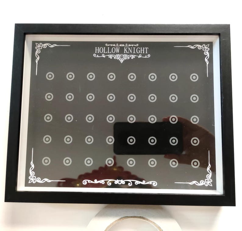 May include: A framed black and white display board with the text "HOLLOW KNIGHT" at the top. The board has a grid of small circles and decorative corner designs. A hand holding a phone is reflected in the display.