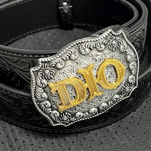 Op de afbeelding: Zwarte leren riem met een decoratieve zilveren gesp. De gesp heeft een goudkleurige "DIO"-inscriptie en sierlijke bloemen- en krulwerkdetails. De riem is opgerold en toont de gesp en het reliëf leer.