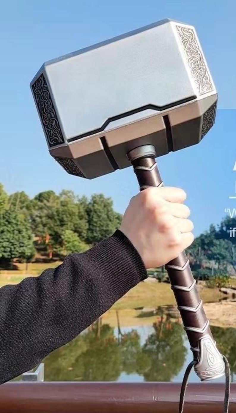 K&ouml;nnte beinhalten: Ein silberner und brauner Requisitenhammer mit aufwendigen Details. Der Hammer hat einen rechteckigen Kopf mit dekorativen Mustern und einen braunen Griff. Der Griff ist mit einem strukturierten Material umwickelt. Am Boden ist eine Lederschlaufe befestigt.