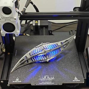 May include: A 3D-printed, silver-colored object with a complex, symmetrical design, illuminated by blue light. The object is on a 3D printer bed, with the printer's components visible in the background. The printer has the text "VORON" on it.