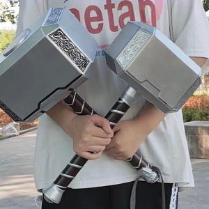 K&ouml;nnte beinhalten: Zwei silberne und schwarze Spielzeugh&auml;mmer mit aufwendigen Designs auf den K&ouml;pfen und dunkelbraunen Griffen. Die H&auml;mmer werden vor einer Person gehalten, die ein wei&szlig;es T-Shirt tr&auml;gt. Die H&auml;mmer scheinen Nachbildungen einer fiktiven Waffe zu sein.
