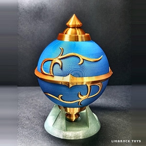 Op de afbeelding: Een bolvormig blauw object met een puntige bovenkant en basis van goudkleurig metaal, voorzien van complexe gouden filigraanpatronen rond het midden. Het object rust op een lichtgrijze standaard met meerdere poten, met de tekst LIONROCK TOYS.