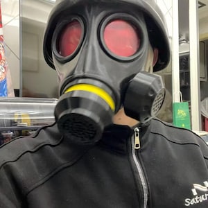 May include: A black gas mask with red lenses and a yellow accent on the filter. The mask is worn with a black zip-up jacket. The text "To the world stage You saturate" is printed on the jacket.
