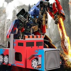 May include: A red, blue, and white cardboard toy truck with the Transformers logo. The truck has a detailed design with wheels, a smokestack, and a front grill. A large image of Optimus Prime is in the background.