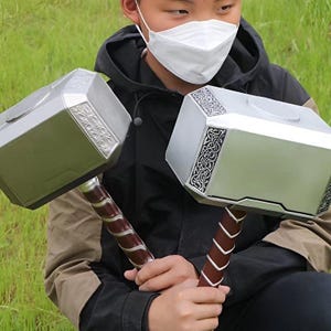 K&ouml;nnte beinhalten: Zwei silberne Spielzeugh&auml;mmer mit braunen Griffen. Die Hammerk&ouml;pfe haben dekorative silberne Muster. Die Griffe sind mit einem braunen Material mit silbernen Akzenten umwickelt. Eine Person mit einer schwarzen Jacke h&auml;lt die H&auml;mmer.