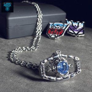 Op de afbeelding: Een zilverkleurige ketting met een blauwe edelsteen hanger. Twee metalen pins met rode, witte, blauwe en paarse ontwerpen zijn ook zichtbaar. Het Lion Rock Toys logo staat in de linkerbovenhoek.