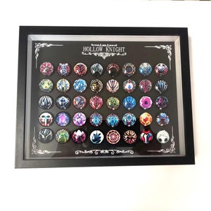 May include: A framed display featuring a collection of colorful, round pins with intricate designs. The pins are arranged in rows against a black background, with the text "HOLLOW KNIGHT" at the top. The frame is black.