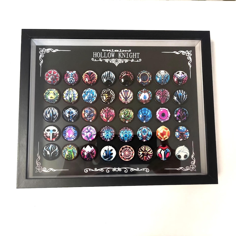 May include: A framed display featuring a collection of colorful, round pins with intricate designs. The pins are arranged in rows against a black background, with the text "HOLLOW KNIGHT" at the top. The frame is black.