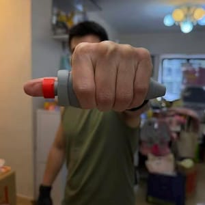 May include: A gray and red finger exerciser. The device has a gray handle and a red ring that fits around the finger. The exerciser is designed to improve grip strength and finger dexterity.