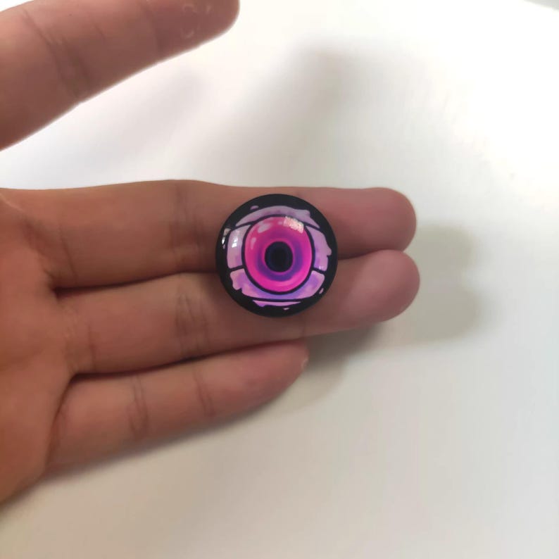 May include: A round, black button with a stylized eye design. The eye features a vibrant pink iris and pupil, surrounded by a lighter pink and purple outline. The button is held in a hand.