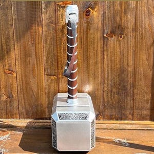 K&ouml;nnte beinhalten: Eine silber-braune Nachbildung von Thors Hammer, Mjolnir. Der Hammer hat einen silbernen Kopf mit dekorativen Mustern und einen braunen Griff mit einem Lederriemen. Der Griff ist spiralf&ouml;rmig gewickelt.