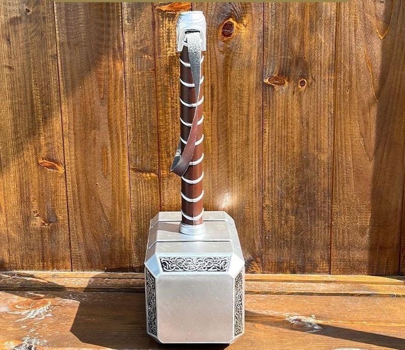 K&ouml;nnte beinhalten: Eine silber-braune Nachbildung von Thors Hammer, Mjolnir. Der Hammer hat einen silbernen Kopf mit dekorativen Mustern und einen braunen Griff mit einem Lederriemen. Der Griff ist spiralf&ouml;rmig gewickelt.