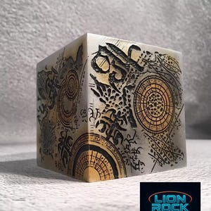 May include: A square cube with a metallic appearance, covered in detailed etchings in black and gold. The design includes circular and linear patterns. The bottom right corner features the "LION ROCK TOYS" logo.