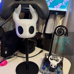Op de afbeelding: Een witte en zwarte gamecontrollerhouder met een gestileerd karakterontwerp. De houder is gemonteerd op een zwarte standaard met een cirkelvormige basis. Een grijze gamecontroller rust erbovenop. Er is ook een kleine decoratieve scène aanwezig.