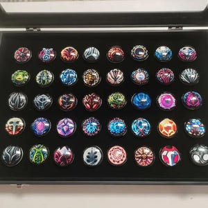 Puede incluir: Una vitrina negra contiene una colección de botones decorativos. Los botones presentan diversos diseños coloridos, incluyendo patrones geométricos y motivos florales. Los botones redondos tienen un acabado brillante. La vitrina está abierta.