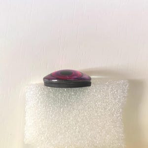 May include: A decorative bottle stopper with a black base and a domed top featuring concentric circles of pink, purple, and black. The stopper is resting on a white, textured surface.