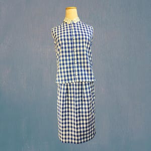 Op de afbeelding: Een blauw en wit geruit mouwloos top en rok set. De top heeft een witte kraag en knopen aan de voorkant. De rok is knielang en heeft een bijpassend ruitpatroon. De outfit wordt getoond op een mannequin.