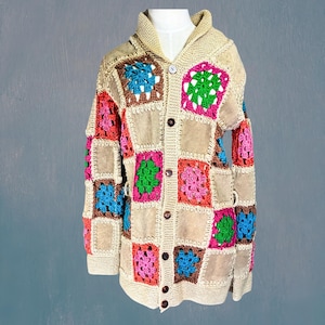 以下が含まれることがあります： ベージュのパッチワークカーディガン、カラフルなかぎ針編みの正方形付き。 ショールカラー、ボタン留め、スエードやクロシェなど、さまざまな素材を使用しています。 かぎ針編みの正方形は、ピンク、グリーン、ブルー、ブラウンの色合いです。