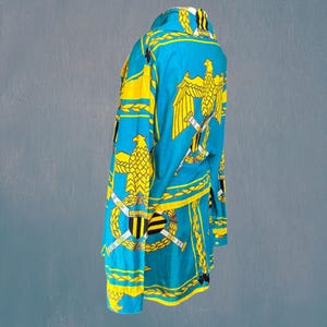 以下が含まれることがあります： 鮮やかなターコイズ色の長袖シャツ。黄色いワシ、剣、月桂樹の繰り返し模様が特徴です。クラシックな襟とボタンダウンのデザインで、装飾的な要素が大胆で人目を引く魅力を加えています。