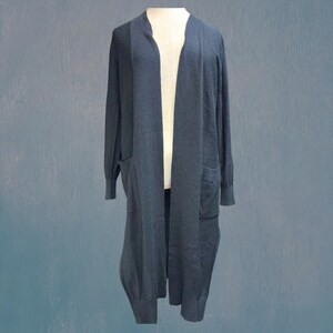 Puede incluir: Un cárdigan largo azul oscuro con dos bolsillos delanteros grandes. El diseño abierto llega hasta los tobillos. El cárdigan se muestra en un maniquí sobre un fondo azul.