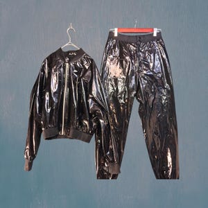 Könnte beinhalten: Ein schwarzes, glänzendes Trainingsanzug-Set, bestehend aus einer Bomberjacke mit silbernem Reißverschluss und passender Hose. Jacke und Hose hängen an separaten Kleiderbügeln vor einem blau-grünen Hintergrund. Das Set scheint aus glänzendem Material zu sein.