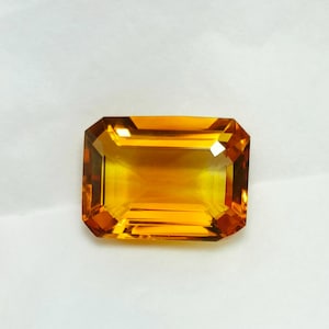 Könnte beinhalten: Ein rechteckig geschliffener, goldgelber Edelstein mit einer brillanten, facettierten Oberfläche. Der Edelstein hat eine warme, strahlende Farbe und eine symmetrische Form, ideal für Schmuckdesign. Der Edelstein ist vor weißem Hintergrund.