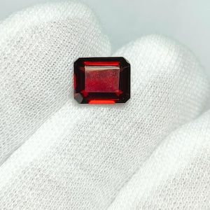 Könnte beinhalten: Ein facettierter, rechteckig geschliffener, tiefroter Edelstein. Der Edelstein hat eine satte, gesättigte Farbe und ist vor einem weißen Hintergrund platziert. Die Kanten des Edelsteins sind scharf definiert und zeigen seine Klarheit und Brillanz.