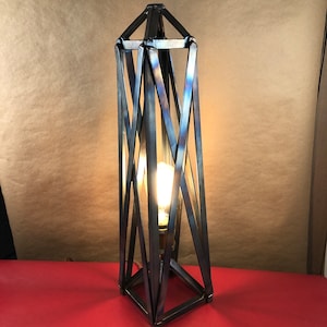 May include: A modern, geometric table lamp with a metal frame and a light bulb. The lamp is made of dark metal and has a criss-cross pattern.