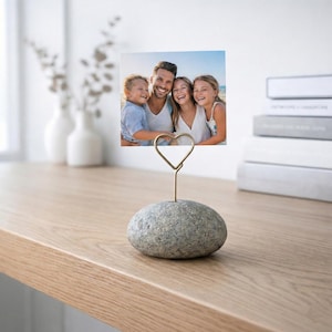 May include: A grey stone photo holder with a gold-coloured wire heart-shaped clip. The holder displays a family photo. The photo holder sits on a light-coloured wooden surface, with white vases and books in the background.