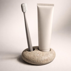 K&ouml;nnte beinhalten: Eine graue Zahnb&uuml;rste mit wei&szlig;en Borsten und eine Tube Zahnpasta befinden sich in einem Steinhalter. Der Halter hat zwei runde &Ouml;ffnungen, eine f&uuml;r die Zahnb&uuml;rste und eine f&uuml;r die Zahnpastatube. Die Zahnpastatube ist wei&szlig;.