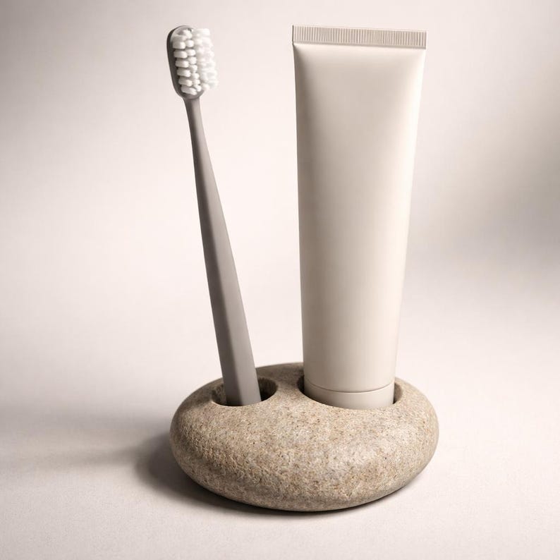 K&ouml;nnte beinhalten: Eine graue Zahnb&uuml;rste mit wei&szlig;en Borsten und eine Tube Zahnpasta befinden sich in einem Steinhalter. Der Halter hat zwei runde &Ouml;ffnungen, eine f&uuml;r die Zahnb&uuml;rste und eine f&uuml;r die Zahnpastatube. Die Zahnpastatube ist wei&szlig;.