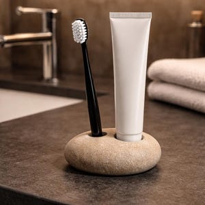 K&ouml;nnte beinhalten: Eine schwarze Zahnb&uuml;rste mit wei&szlig;en Borsten und eine wei&szlig;e Tube Zahnpasta befinden sich in einem beigefarbenen Steinhalter. Der Halter hat zwei kreisf&ouml;rmige &Ouml;ffnungen. Die Artikel stehen auf einer dunklen Arbeitsplatte, mit einem Wasserhahn und Handt&uuml;chern im Hintergrund.