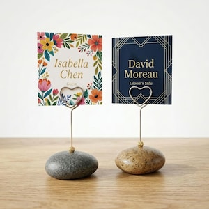 May include: Two place card holders with name cards. One card has a floral design with the name "Isabella Chen" and "Guest". The other card has a geometric design with the name "David Moreau" and "Groom's Side". The holders have a heart-shaped clip and sit on a stone base.