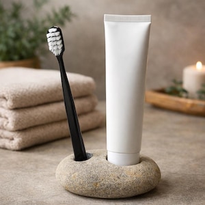 K&ouml;nnte beinhalten: Ein schwarzer Zahnb&uuml;rstenhalter mit wei&szlig;en Borsten und eine wei&szlig;e Tube Zahnpasta werden in einem Steinst&auml;nder pr&auml;sentiert. Der St&auml;nder steht auf einer neutralen Oberfl&auml;che, mit gefalteten Handt&uuml;chern im Hintergrund. Eine brennende Kerze ist sichtbar.